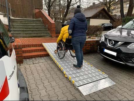 Frau schiebt Rollstuhlfahrer eine mobile Rampe hoch